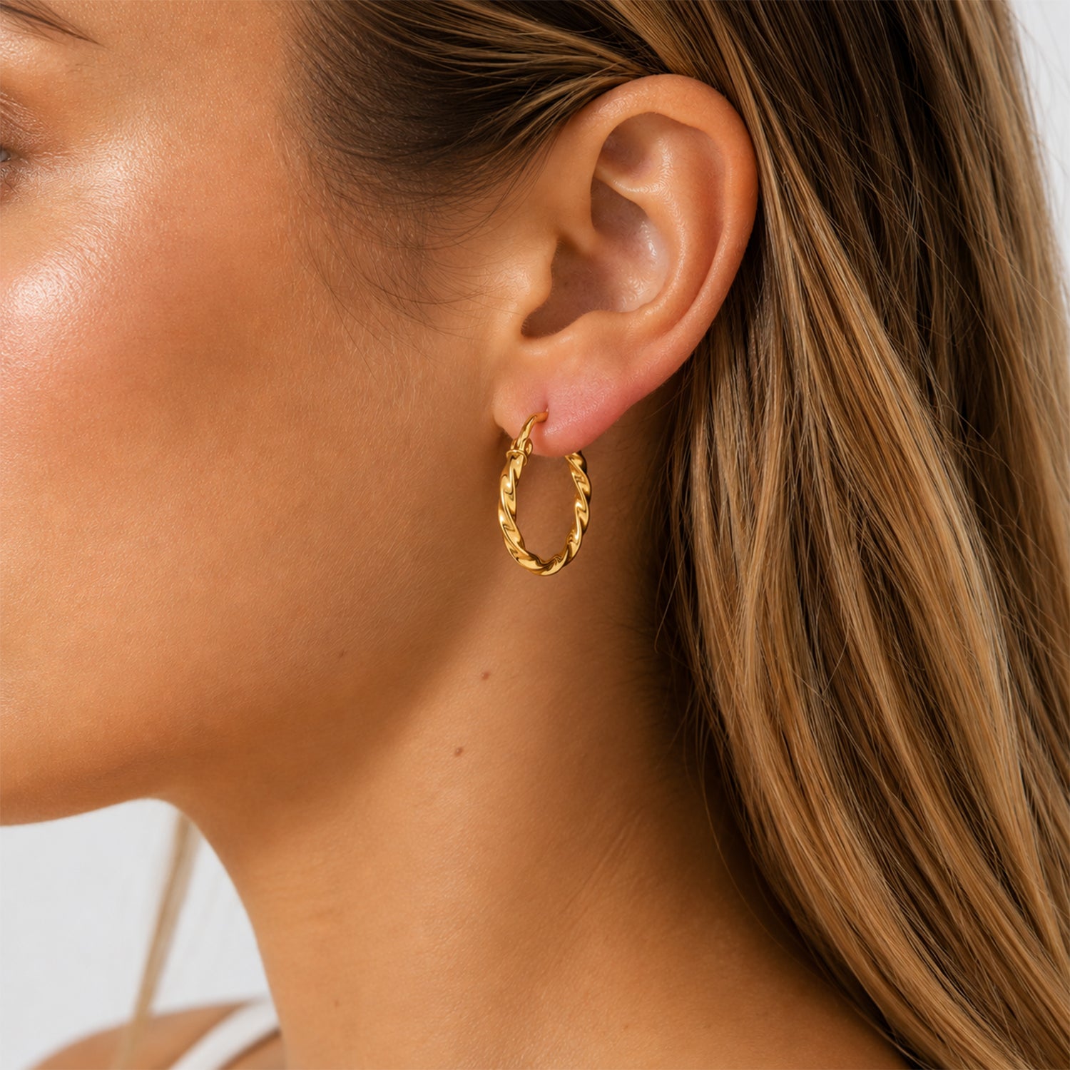 A close-up of a woman's ear wearing a small twisted gold hoop earring.
