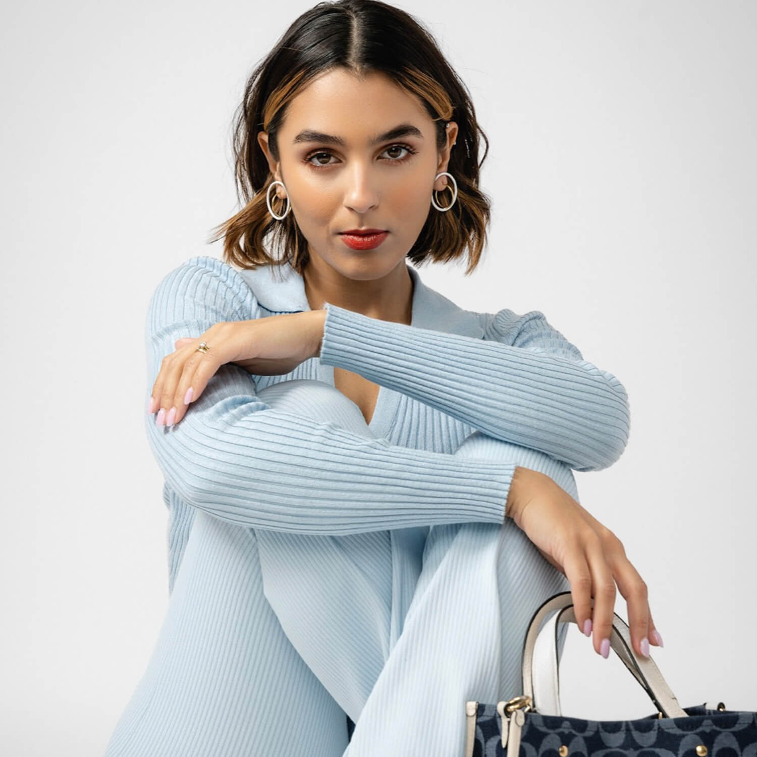 A woman with short brown hair and hoop earrings poses in a light blue ribbed knit cardigan, holding a navy blue patterned handbag with gold hardware against a light gray studio background.