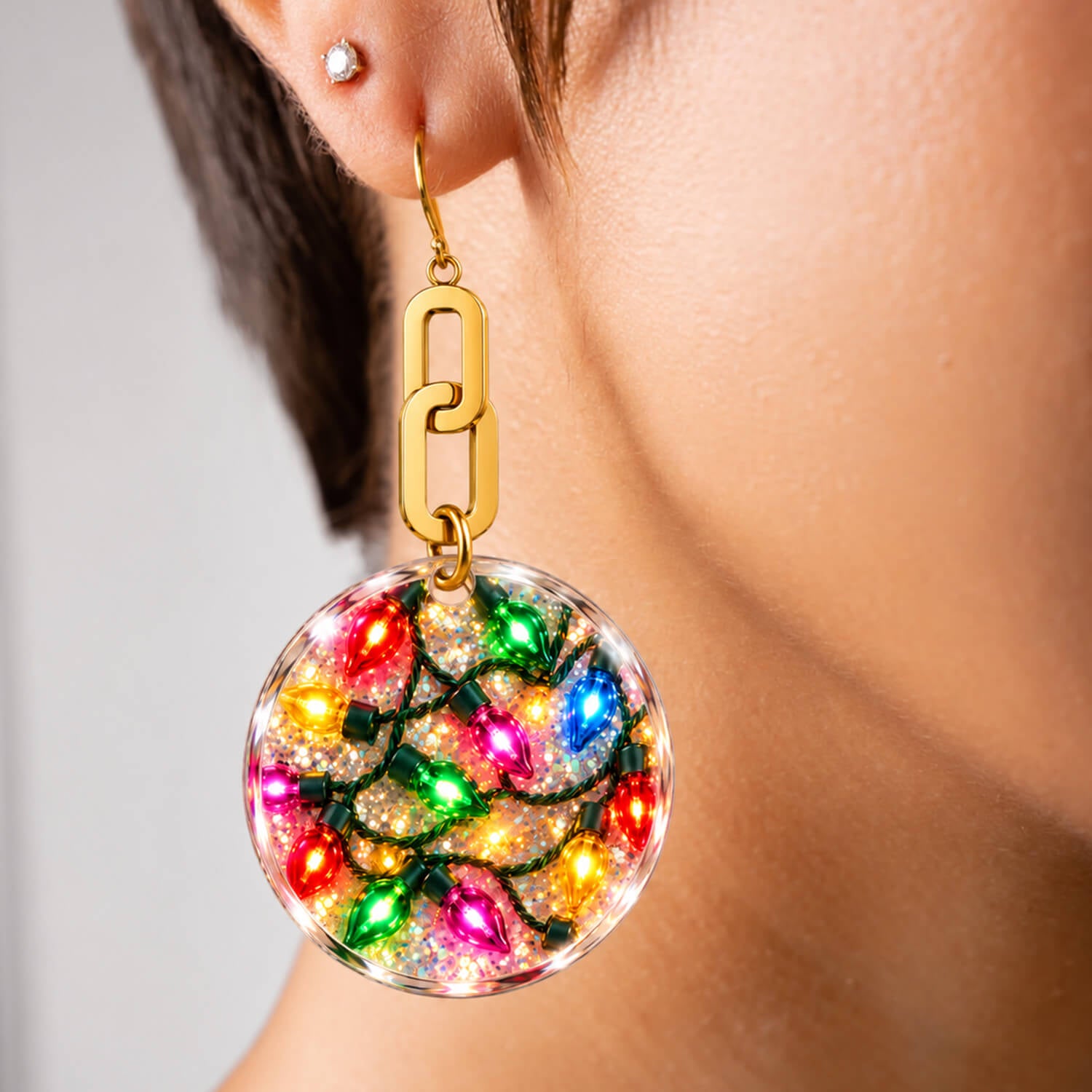 A close-up of a person's ear wearing a dangling earring. The earring has a gold-tone chain-link top and a clear round drop filled with multicolored Christmas light bulbs and glitter.