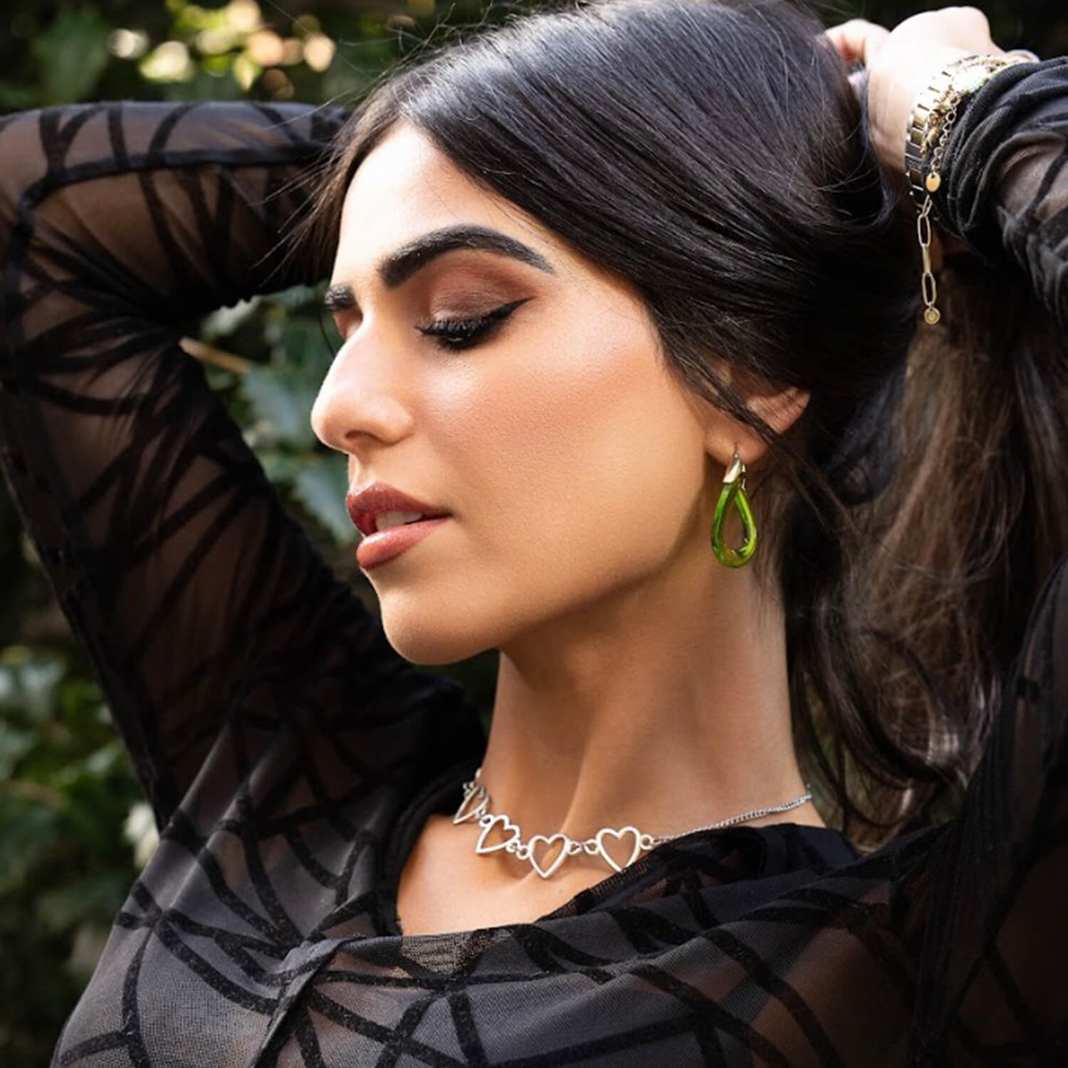 Woman wearing a black sheer top with green earrings and a necklace, outdoors.