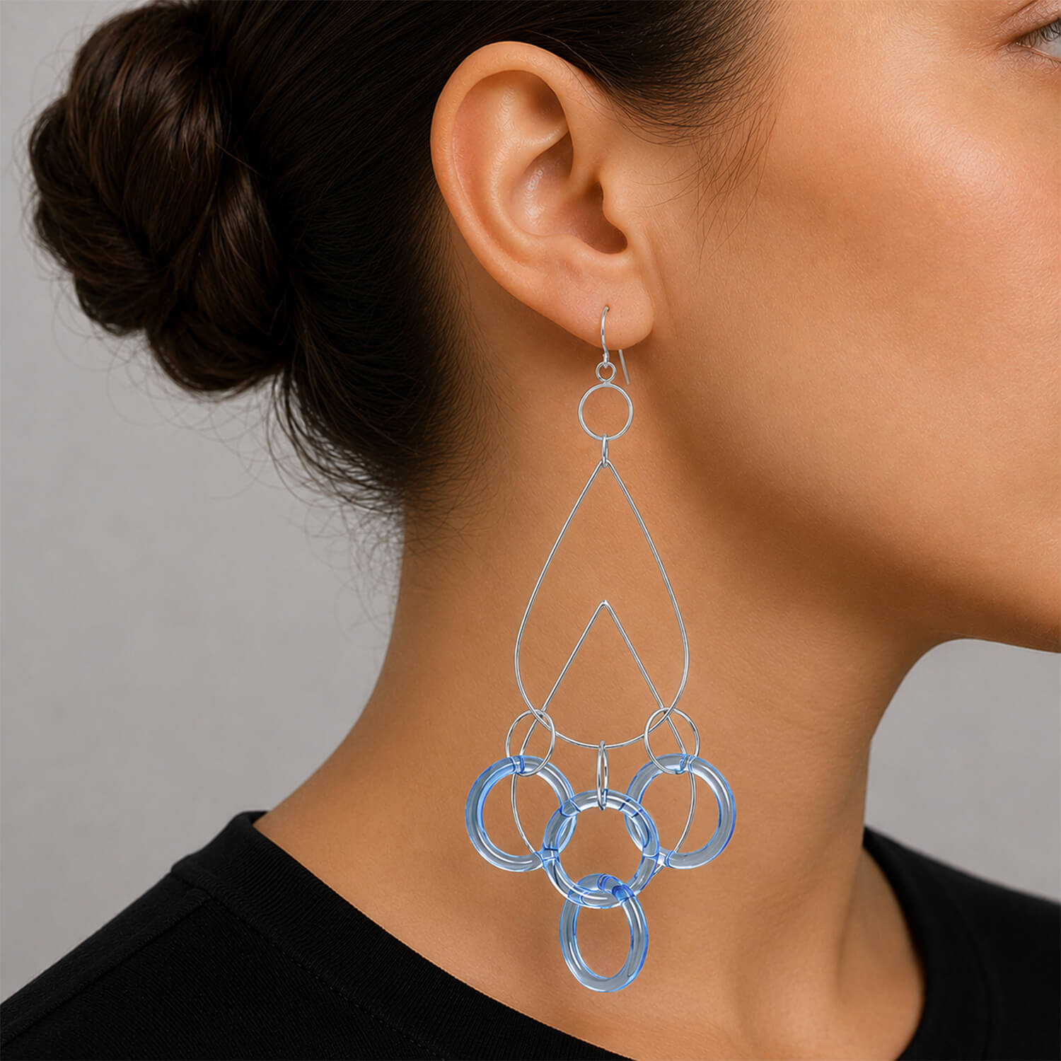 A close-up of a woman's ear and neck wearing a long, geometric dangle earring made of silver-tone metal and translucent light blue rings, against a neutral gray background.