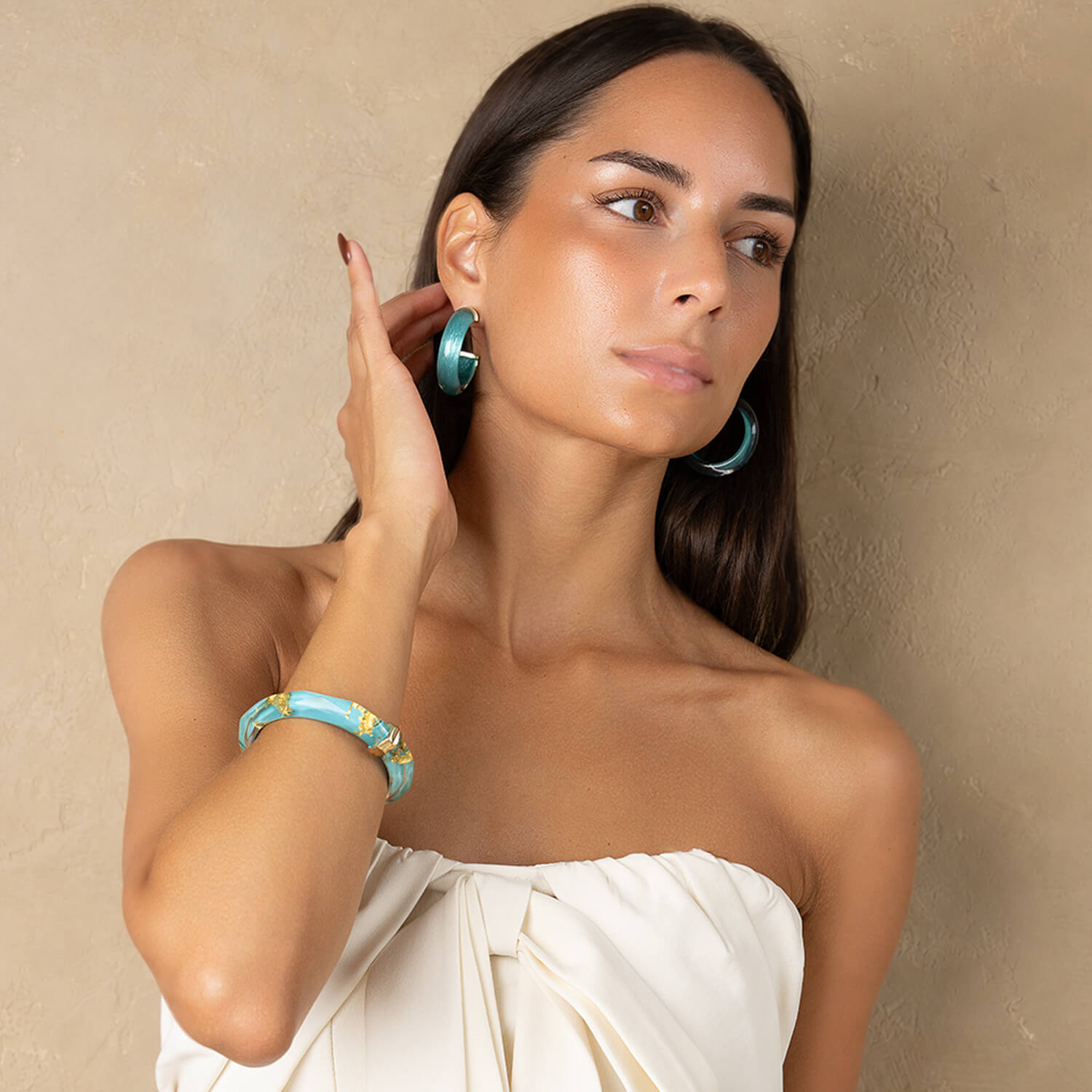 A woman wearing turquoise hoop earrings and a matching turquoise bracelet with gold accents, posing against a textured beige wall.