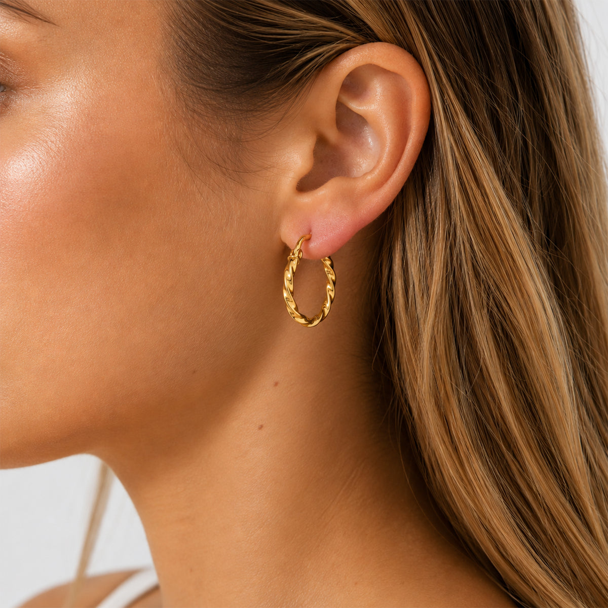 A close-up of a woman's ear wearing a small twisted gold hoop earring.