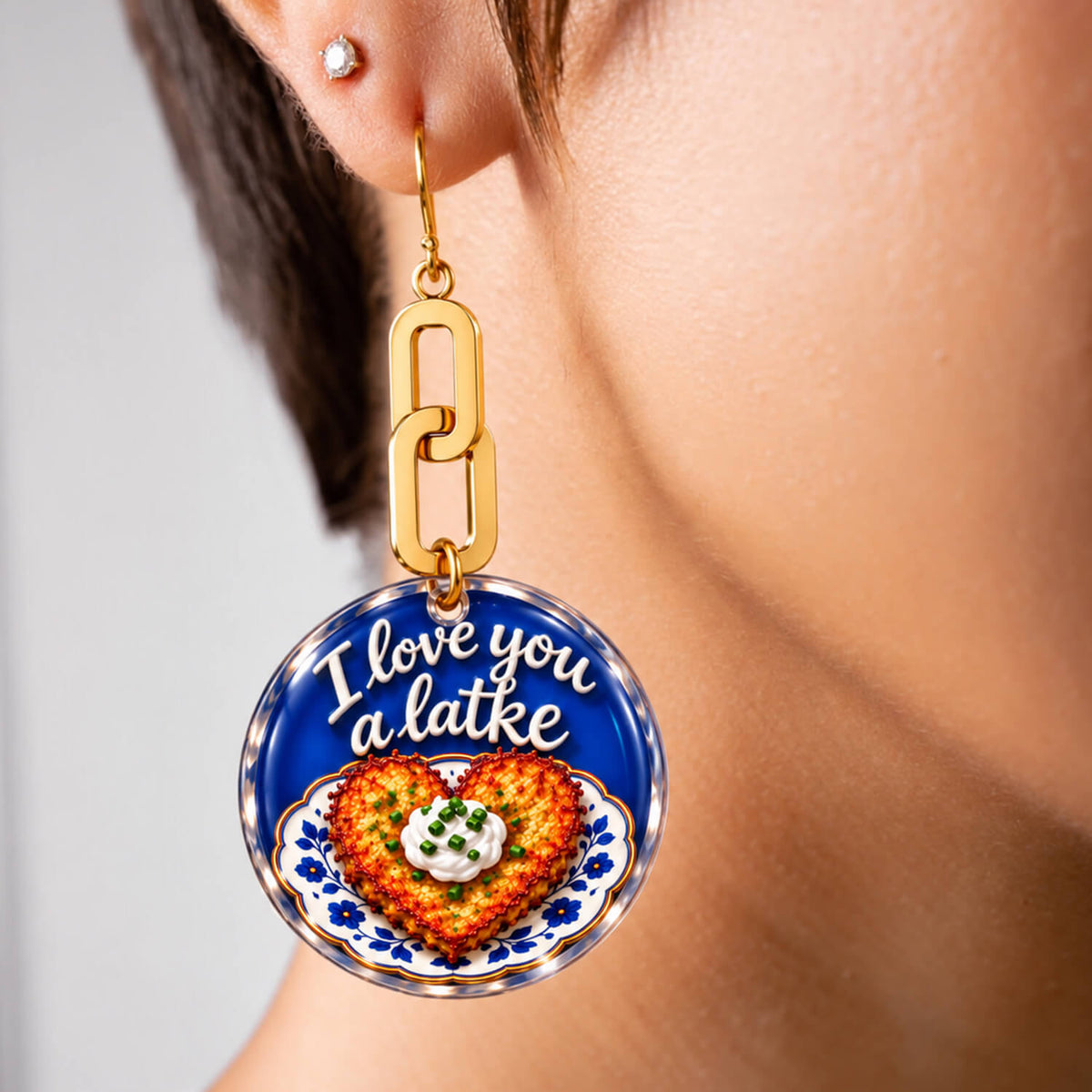 A close-up of a person wearing a gold-tone dangle earring with a round blue charm that reads 'I love you a latke' above a heart-shaped latke with a dollop of cheese. The person's ear, neck, and hair are visible against a soft light gray background.