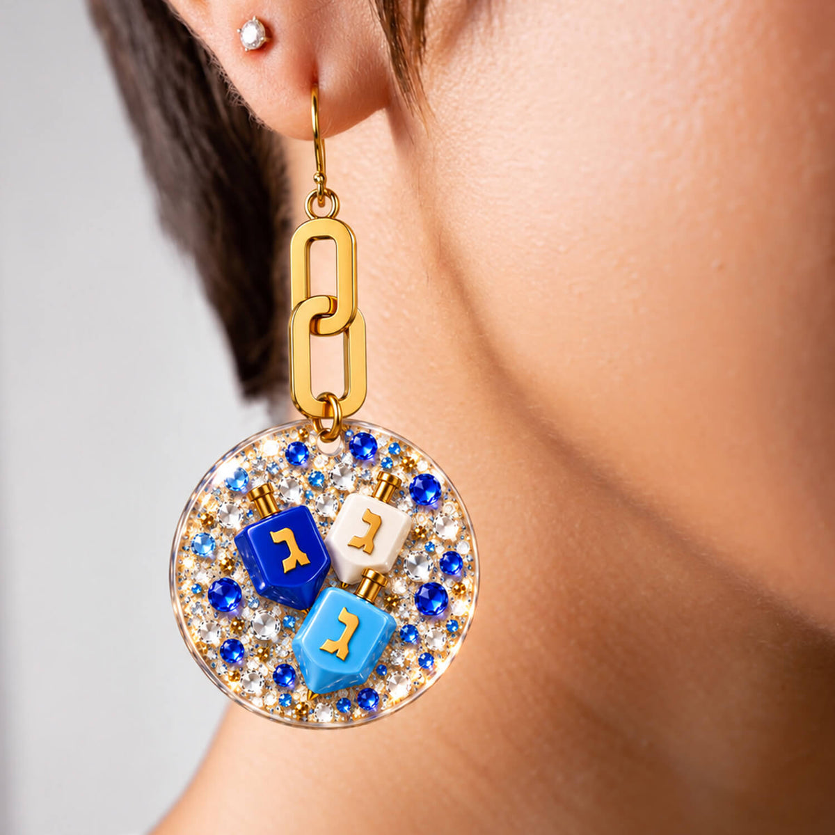 A close-up of a model's ear wearing a gold dangle earring with a circular rhinestone-encrusted disc and three blue, white, and light blue dreidels.