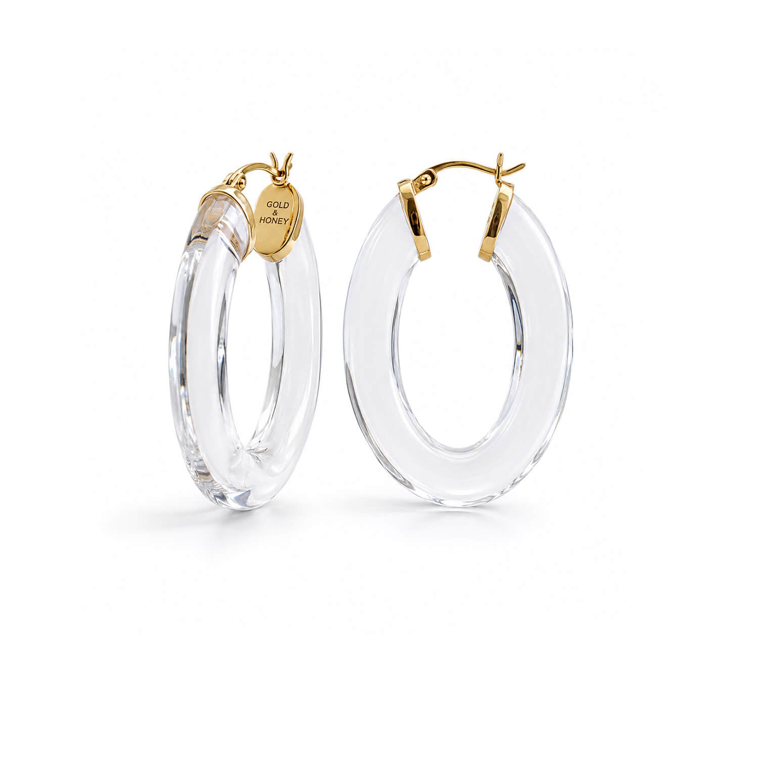 A pair of large oval hoop earrings made of transparent clear acrylic with gold-tone hardware and a small branded tag, shown on a clean white background.