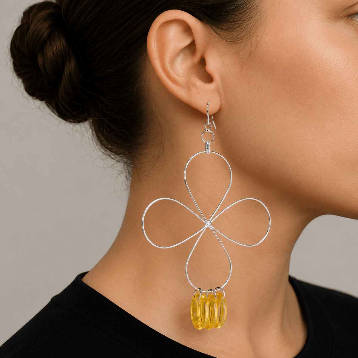 A close-up of a model's ear and neck wearing a large silver wire flower dangle earring with a yellow faceted bead charm, set against a neutral gray background.