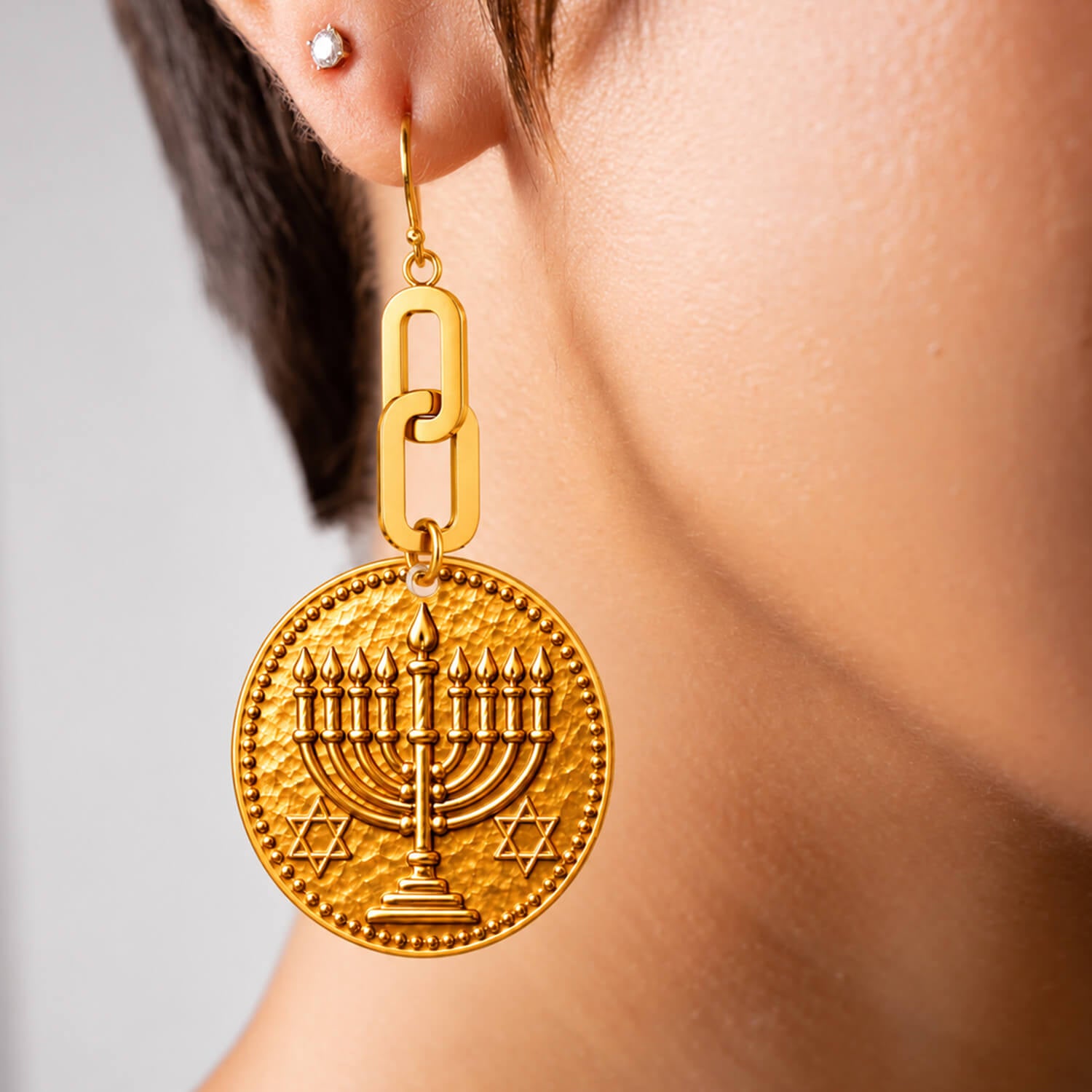 A pair of gold-tone dangle earrings on a white background. One earring features a round pendant with an embossed menorah and Star of David, while the other shows the back of a larger oval pendant with a metallic bronze glitter finish and a gold chain link connector.