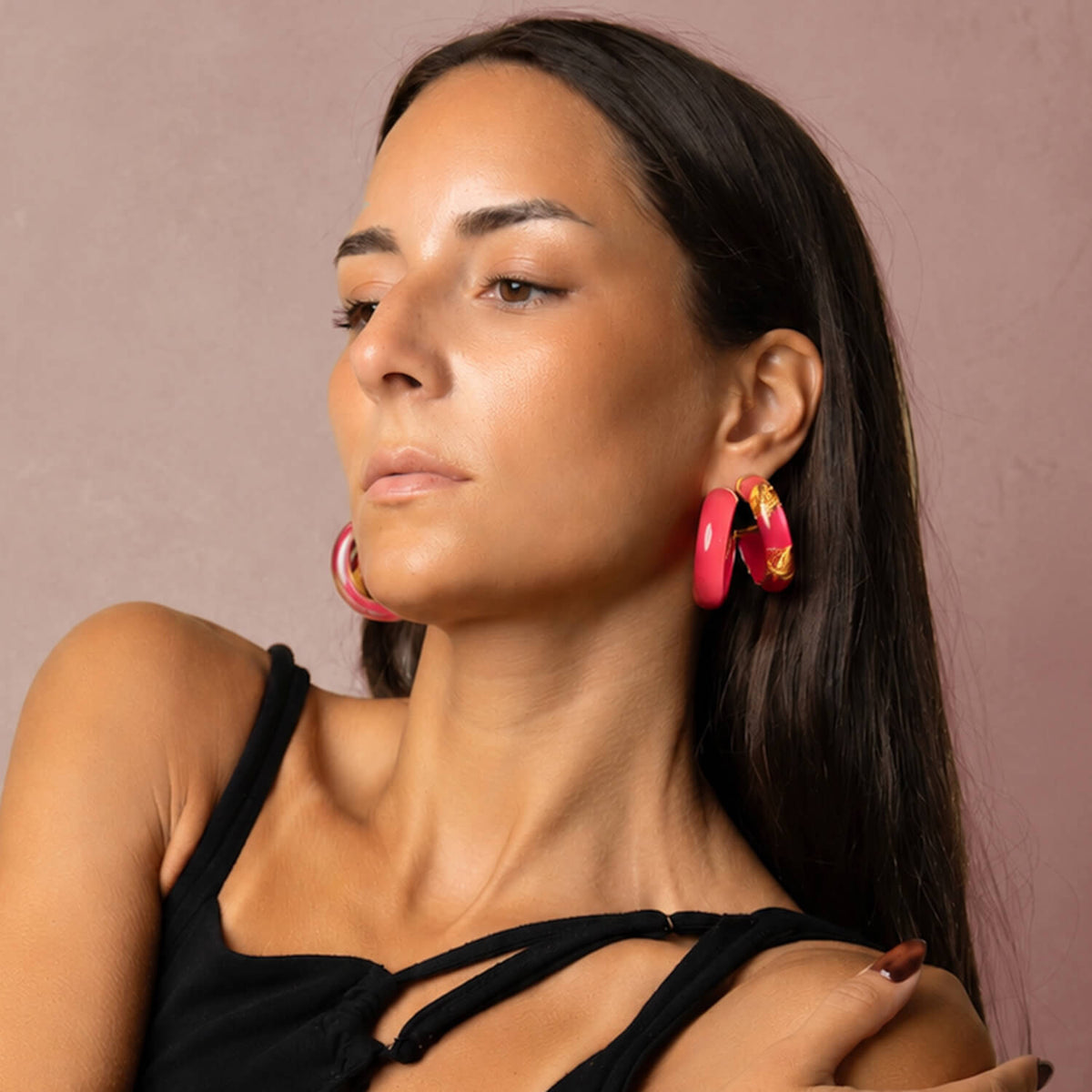 A close-up of a woman wearing vibrant pink hoop earrings with gold accents, paired with a black strappy top against a soft mauve background.