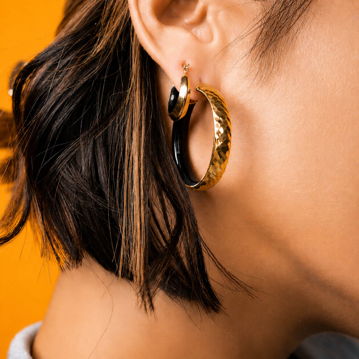 A close-up of a person wearing a pair of large gold hoop earrings, with one earring showing a hammered texture and the other a smooth polished finish. The background is a solid bright orange.