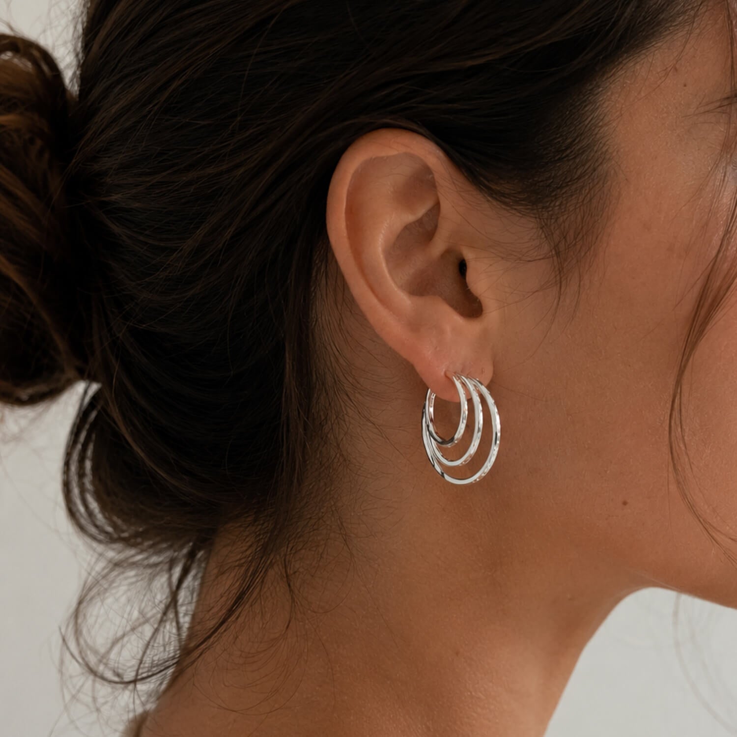 A close-up of a model's ear wearing a pair of silver triple-hoop earrings against a neutral light gray background.