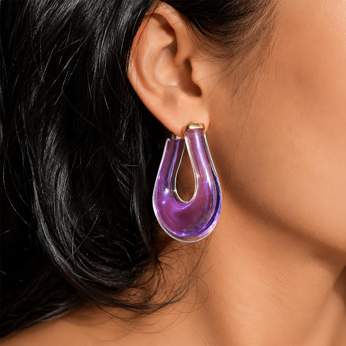 A close-up of a person's ear wearing a large, teardrop-shaped hoop earring made of translucent purple glass with a gold-tone top post.