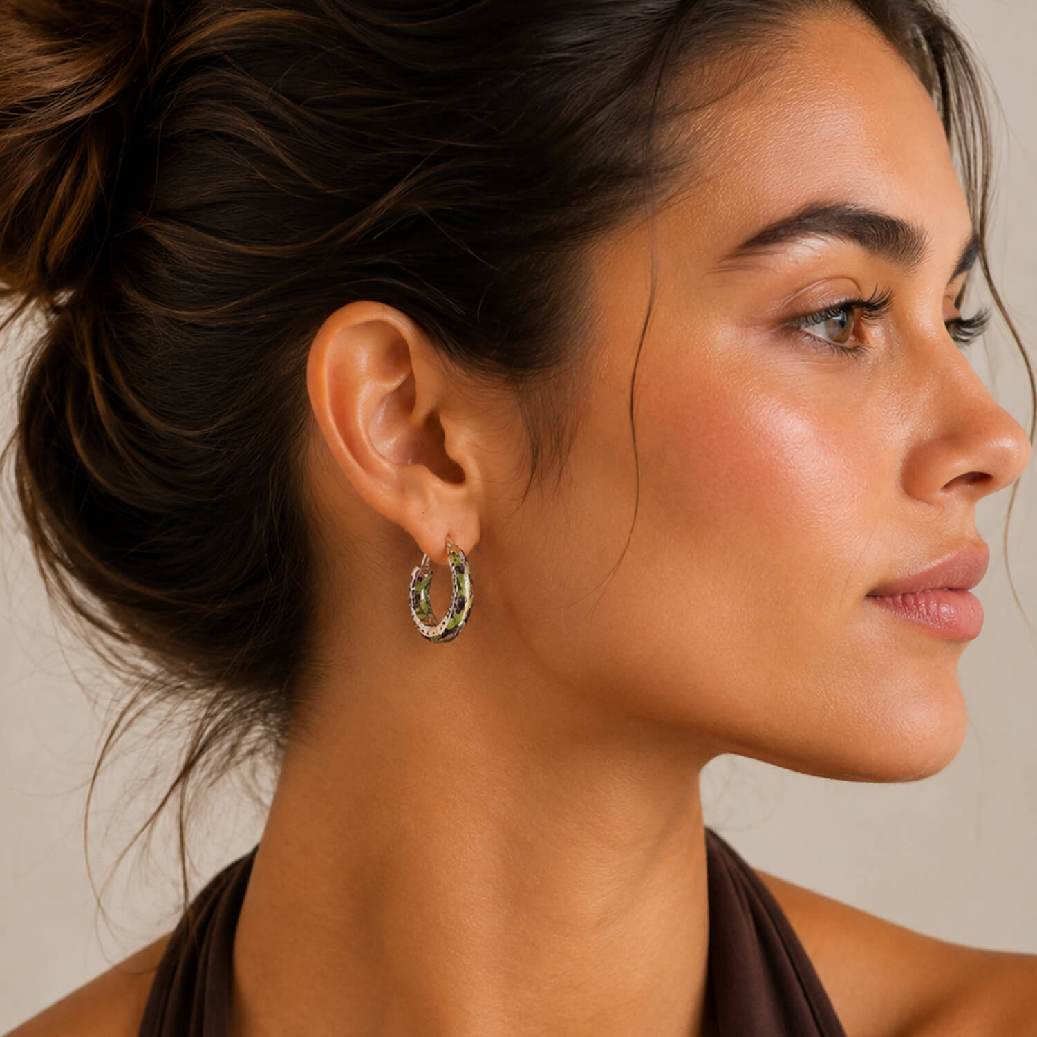 A close-up side profile of a woman wearing a small snake print hoop earring with green and diamond accents against a neutral beige background.