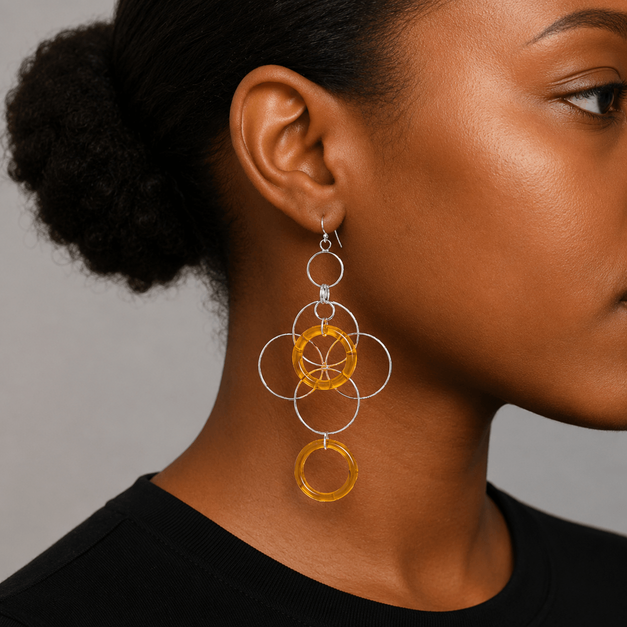 A close-up of a woman wearing a dangling earring. The earring features multiple interlocking silver-tone rings with translucent amber-yellow circular accents, shown against a neutral gray background.