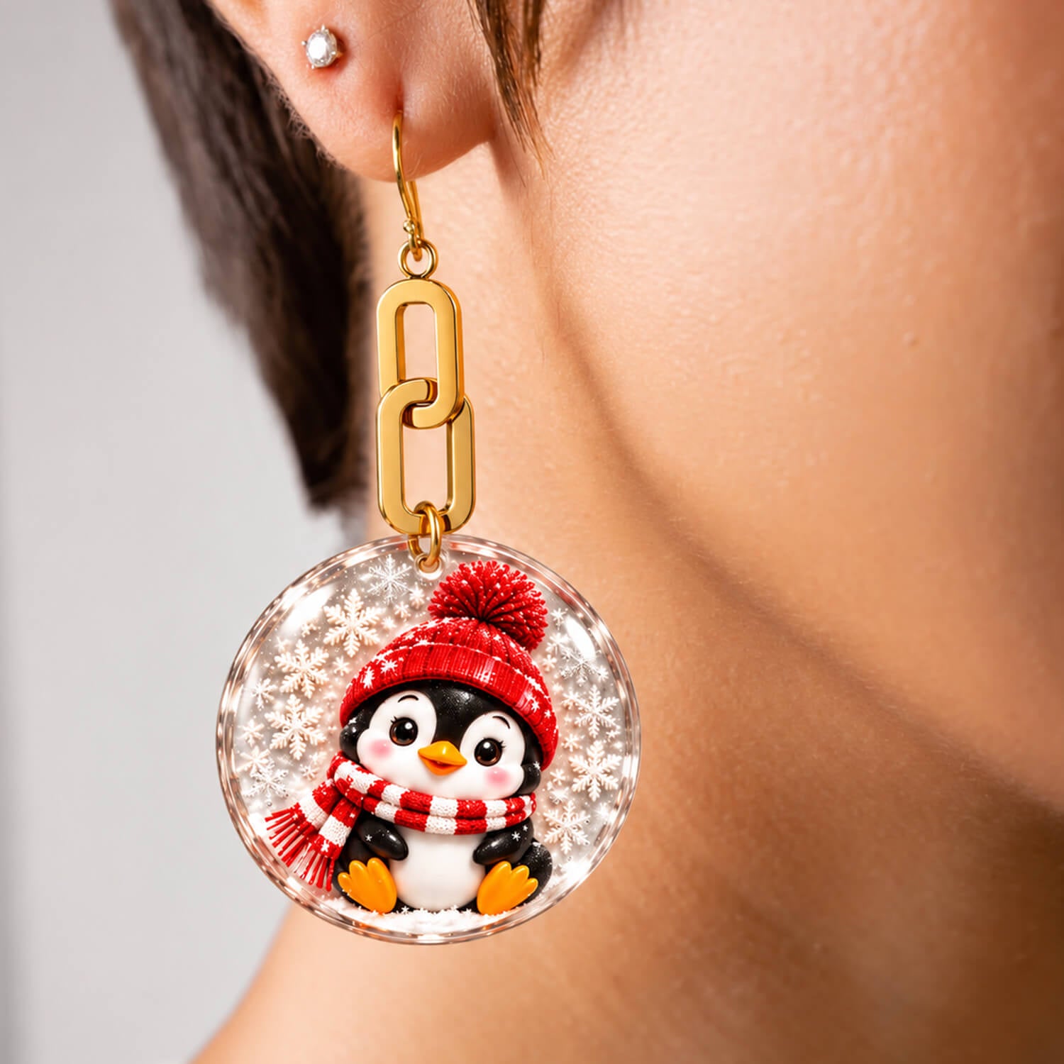 A pair of dangling earrings on a white background, showing both the front and back views. The front view features a round clear resin pendant with a penguin wearing a red knit hat and scarf, suspended from a gold link.