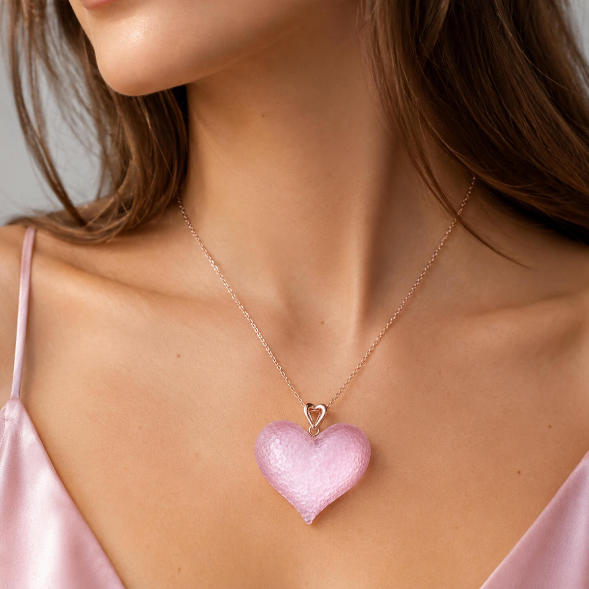 A close-up of a model wearing a delicate rose gold chain necklace with a textured, glittery light pink heart-shaped pendant.