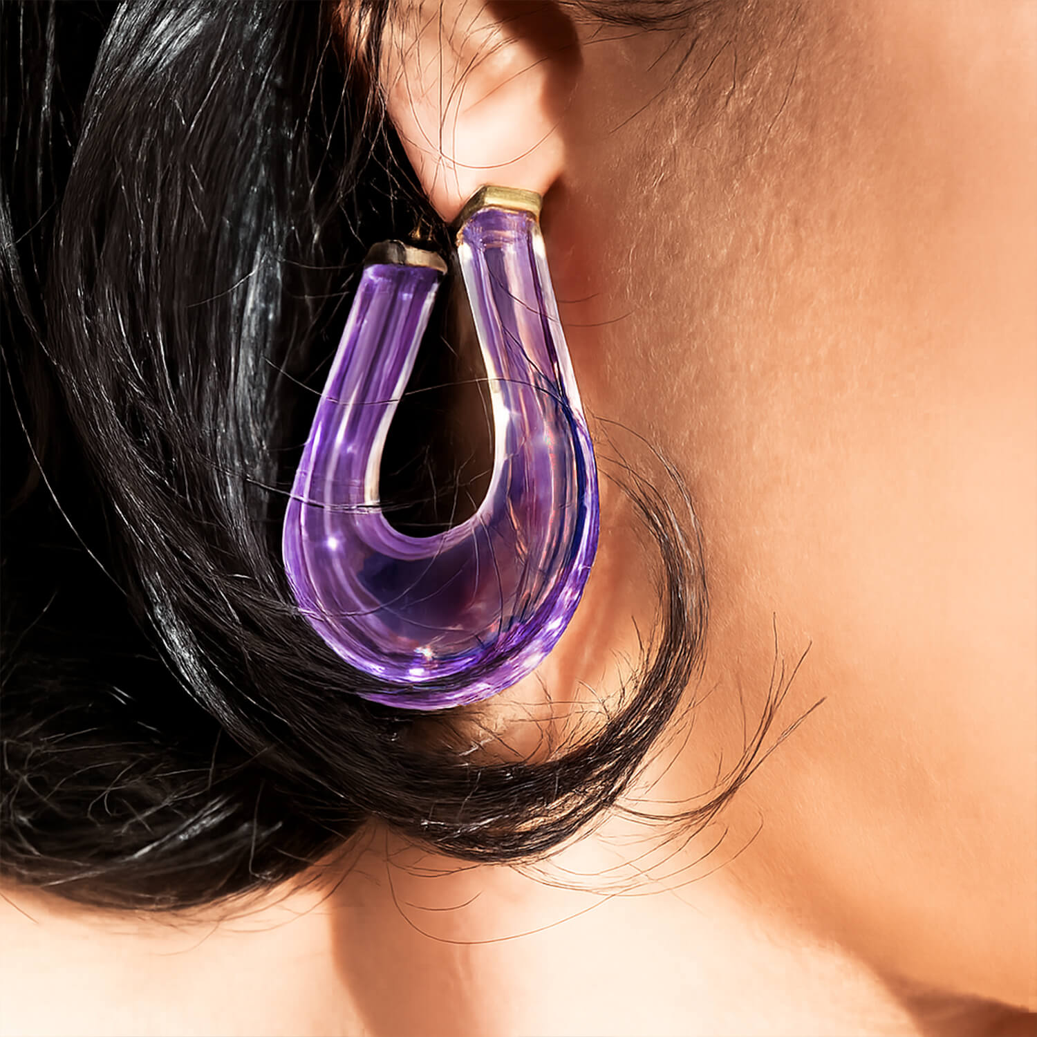 A close-up of a person wearing a large, translucent purple U-shaped hoop earring with gold-toned metal findings, set against warm skin tones and dark hair.