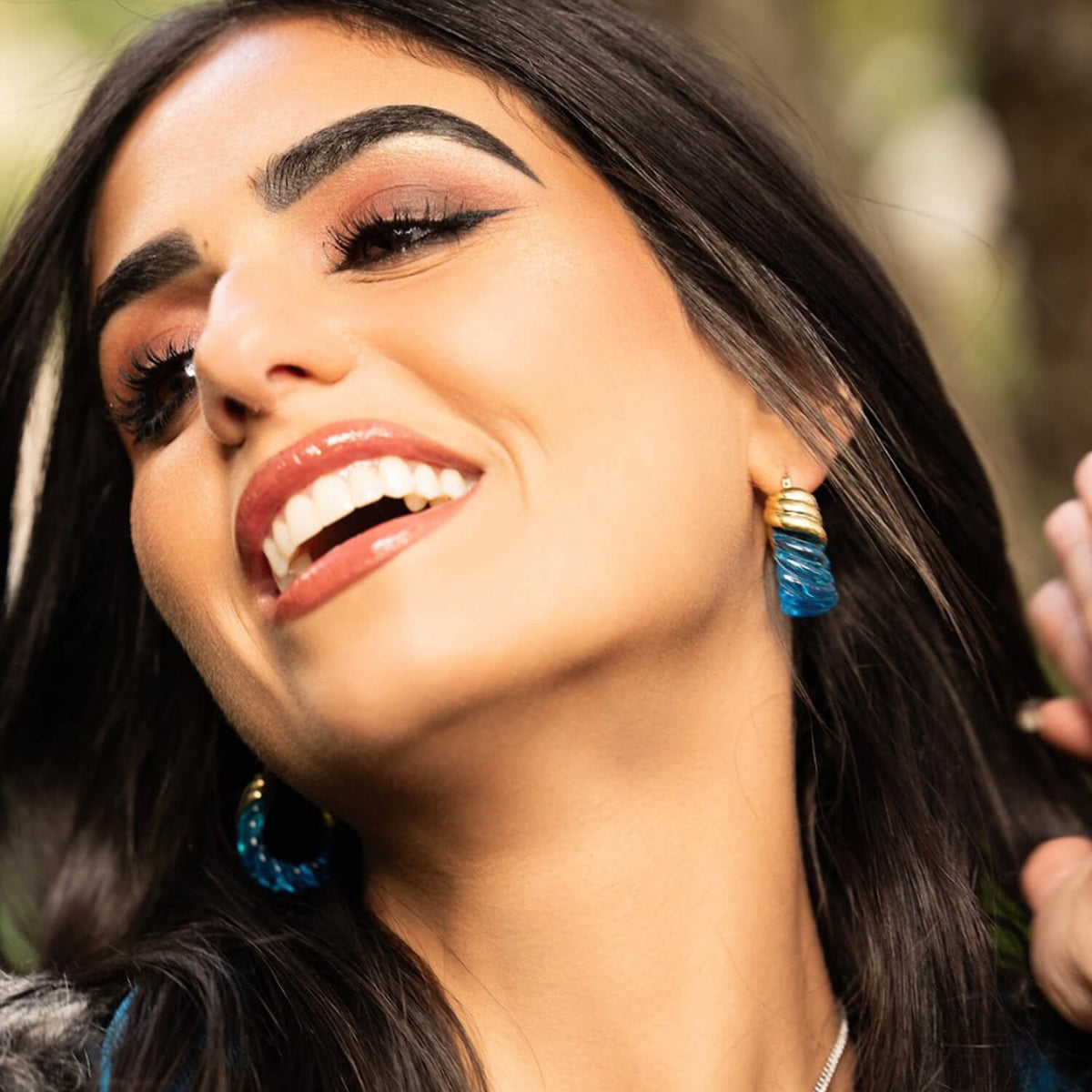 Close-up of a woman with dark hair and blue earrings against a blurred natural background