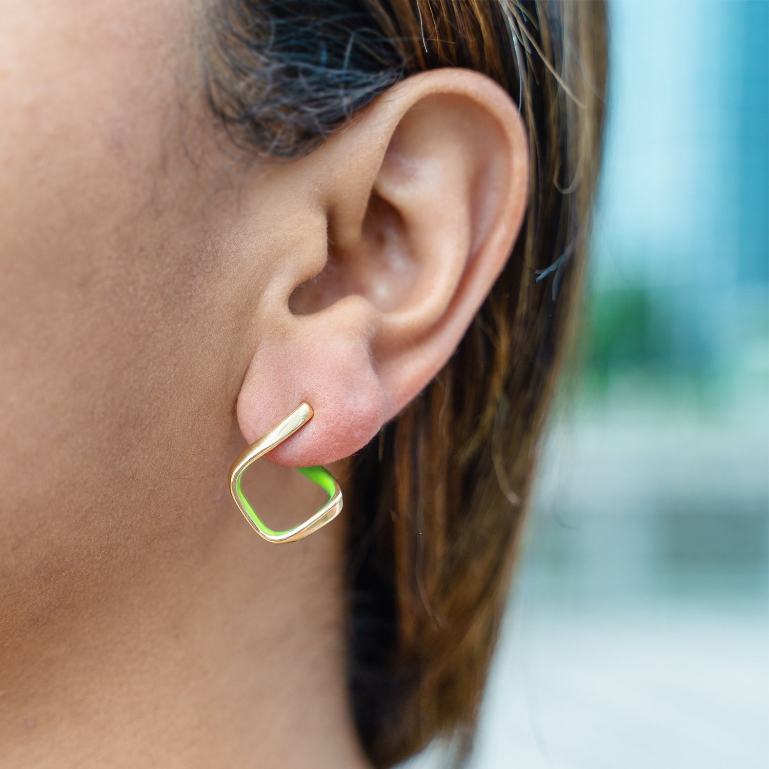A close-up of a person's ear wearing a small gold hoop earring with a bright green inner lining, against a softly blurred outdoor background.