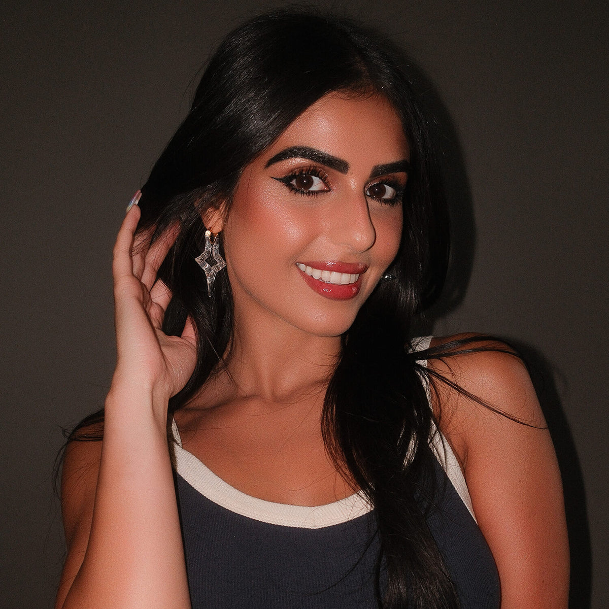 A woman with long black hair and dramatic eye makeup is shown in a close-up portrait, wearing a sparkling clear star earring against a dark gray background.