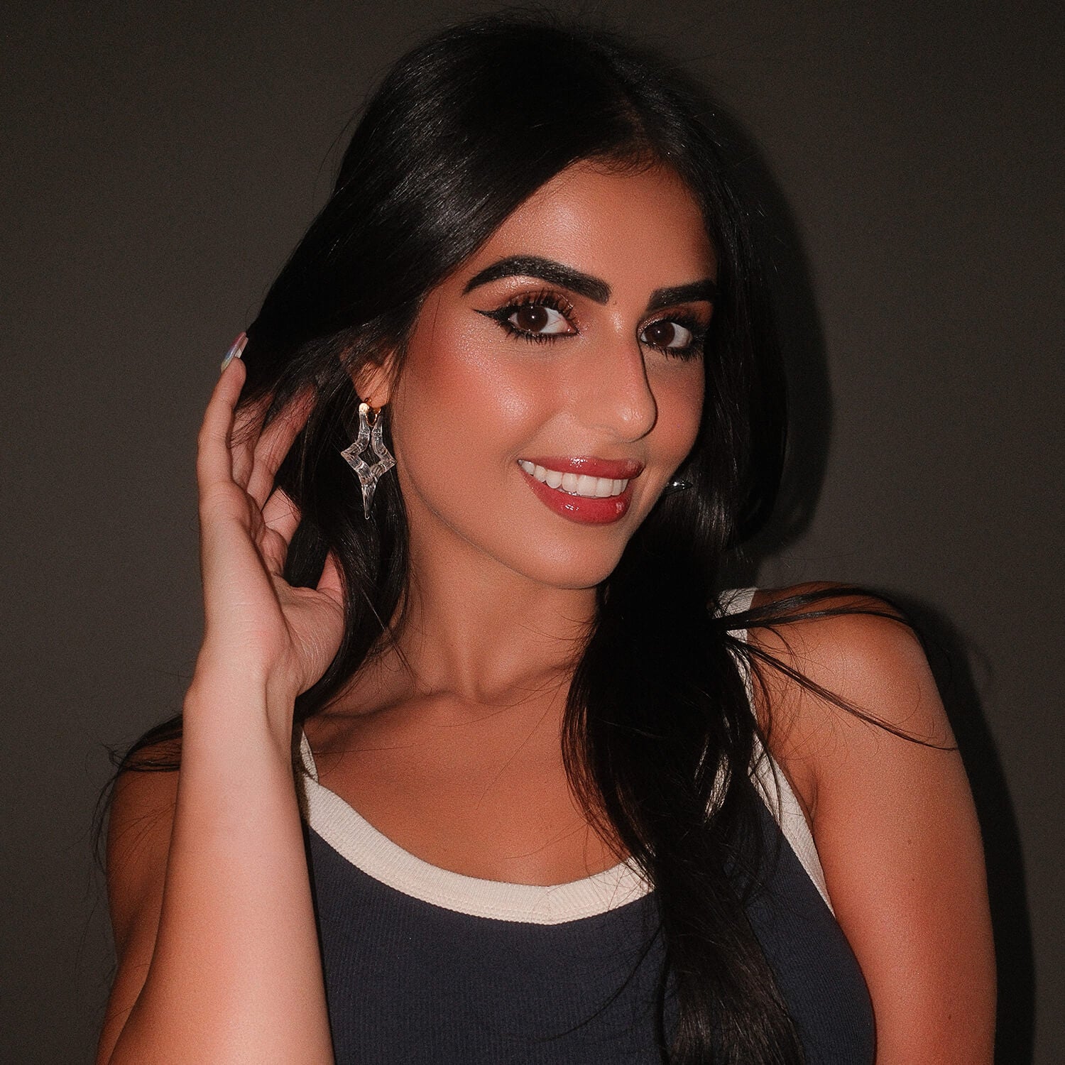 A woman with long black hair and dramatic eye makeup is shown in a close-up portrait, wearing a sparkling clear star earring against a dark gray background.