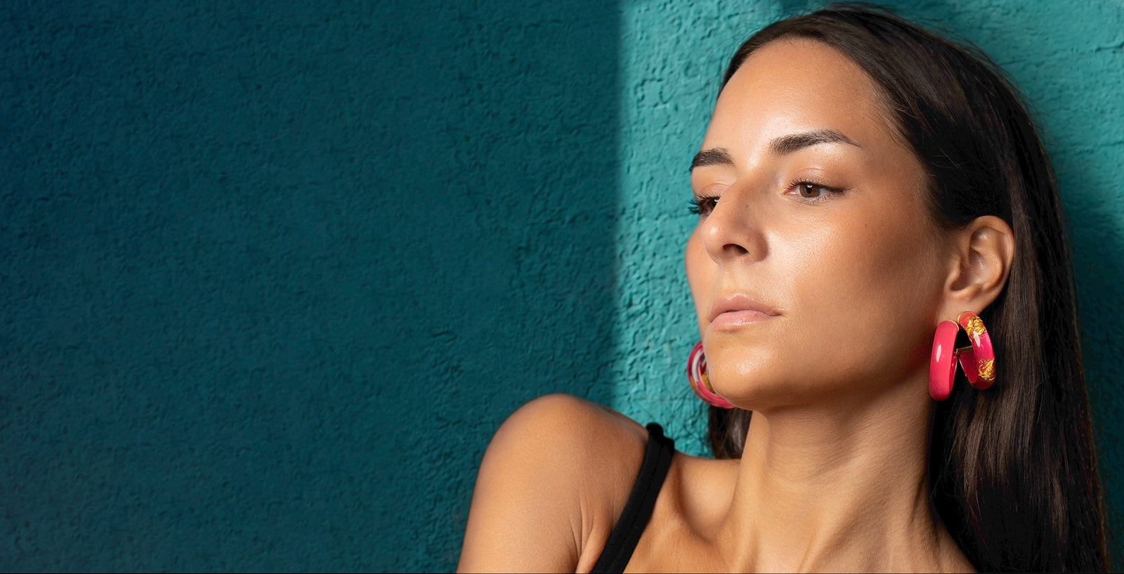A woman with dark hair is shown in profile against a textured teal wall, wearing large bright pink hoop earrings with gold inner detailing.