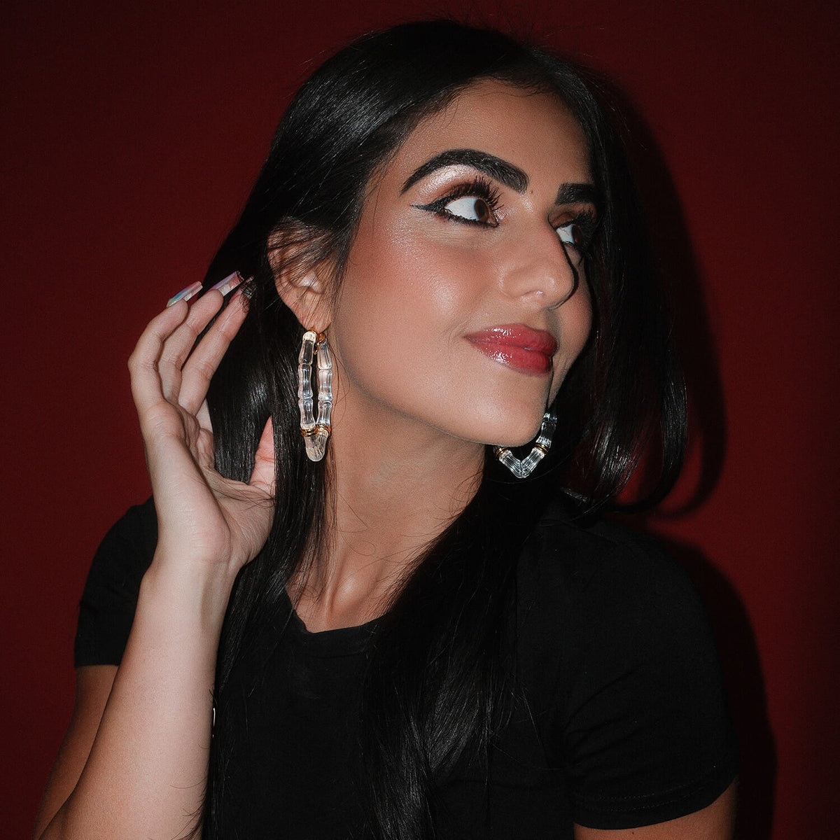 A woman with long black hair and winged eyeliner wearing large clear and gold hoop earrings posing against a dark red background.